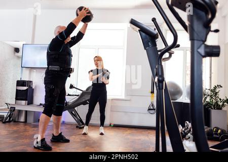 Ein älterer Mann im ems-Anzug trainiert mit einem persönlichen Fitnesstrainer in einem modernen, hellen Fitnessstudio. Ein alter männlicher Sportler, der Sport mit gewichtetem Ball macht Stockfoto