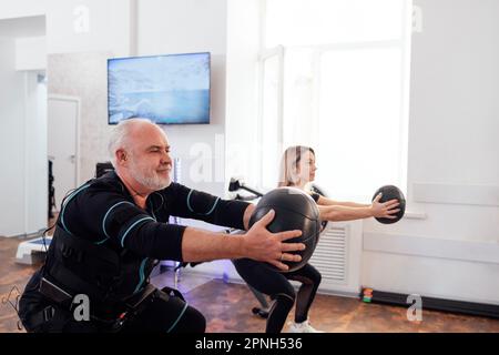 Der grauhaarige ältere Mann im ems-Anzug trainiert mit einem persönlichen Fitnesstrainer in einem modernen Fitnessraum. Alte männliche Sportlerbesetzungen mit gewichtetem Ball bei Functional e Stockfoto