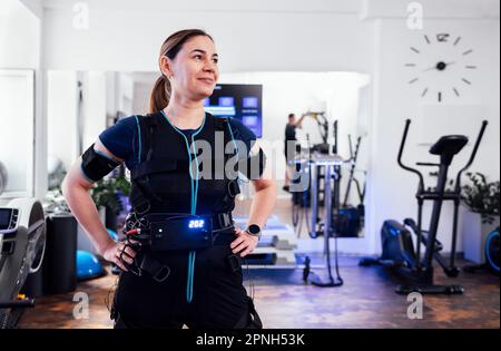 Junge lächelnde Frau, die sich in einem elektrischen Muskelstimulationskostüm im Fitnessclub oder Gym posiert. Selbstbewusste Sportlerin im rettungssanitäter-Anzug mit den Händen an ihrem Bell Stockfoto