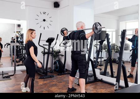 Ein älterer Mann im rettungssanitäter-Anzug trainiert mit einem persönlichen Trainer im Fitnessstudio oder Fitnessclub. Grauhaarige Sportsmann-Squats mit Glocke bei elektrischem Muskelreiz Stockfoto