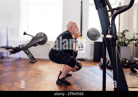 Ein älterer Mann im rettungssanitäter-Anzug trainiert mit einem persönlichen Trainer im Fitnessstudio oder Fitnessstudio. Alte Sportlerbesetzungen mit gewichtetem Ball bei elektrischer Muskelreizung Stockfoto