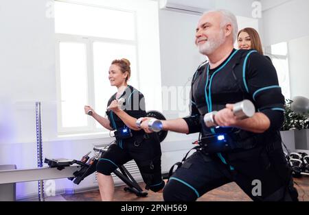 Ein paar lächelnde ältere Menschen in notarztanzügen, die im Fitnessraum mit Kurzhanteln trainieren. Grauhaariger Mann und dicke Frau trainieren mit persönlichem T-Shirt Stockfoto