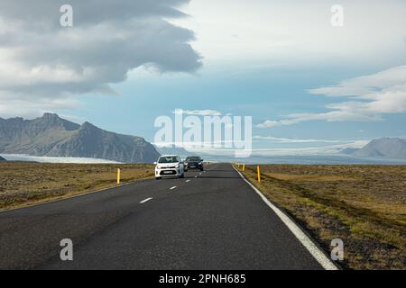 Island - August 2021: suv-Autos auf einer isländischen Straße mit wunderschöner grüner Landschaft, schneebedeckten Bergen und Vatnajokull-Gletscher im Hintergrund Stockfoto