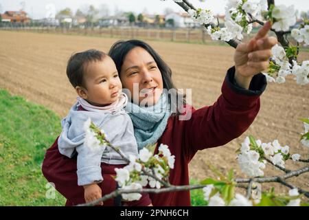 Nahaufnahme einer lächelnden Mutter und Babytochter, die während der Blütenzeit im Frühling Kirschblüten sammeln. Mutterschaftskonzept und Zeitvergnügen Stockfoto