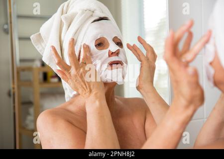 Reife, schöne Frau, die die Gesichtsmaske abnimmt. In den Spiegel im Bad, in ein Handtuch gewickelt. Hautpflegeroutine und Anti-Aging-Behandlung Stockfoto