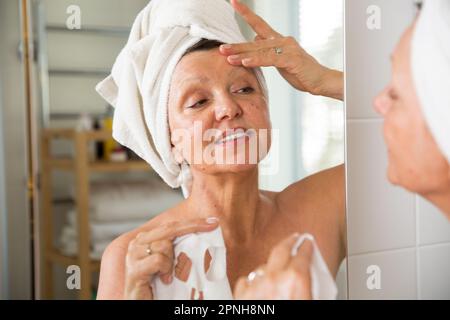 Reife, schöne Frau, die die Gesichtsmaske abnimmt. In den Spiegel im Bad, in ein Handtuch gewickelt. Hautpflegeroutine und Anti-Aging-Behandlung Stockfoto