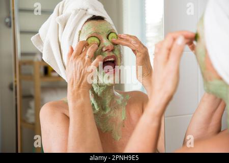 Reife, schöne Frau, die eine grüne hausgemachte Heimwerker-Gesichtsmaske auf der Haut aufträgt. Im Bad in den Spiegel schauen, Spaß haben. In ein Handtuch gewickelt. Hautpflege Stockfoto