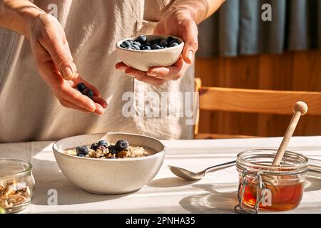 Frau bereitet eine gesunde Ernährung zu, veganes, nahrhaftes Frühstück. Weibliche Hand, die Blaubeeren in die Schüssel mit Haferbrei mit Walnüssen und Honig schüttet. Stockfoto