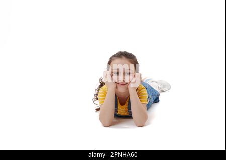 Kaukasisches kleines Mädchen in gelbem T-Shirt und Jeansanzug, auf dem Bauch liegend, in die Kamera schauend, isolierter weißer Hintergrund Stockfoto