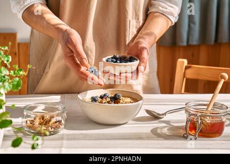 Frau bereitet eine gesunde Ernährung zu, veganes, nahrhaftes Frühstück. Weibliche Hand, die Blaubeeren in die Schüssel mit Haferbrei mit Walnüssen und Honig schüttet. Stockfoto