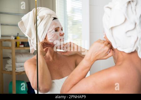 Reife, schöne Frau, die eine Gesichtsmaske auf der Haut aufträgt. In den Spiegel im Bad, in ein Handtuch gewickelt. Hautpflegeroutine und Anti-Aging Stockfoto