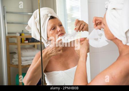 Reife, schöne Frau, die eine Gesichtsmaske auf der Haut aufträgt. In den Spiegel im Bad, in ein Handtuch gewickelt. Hautpflegeroutine und Anti-Aging Stockfoto
