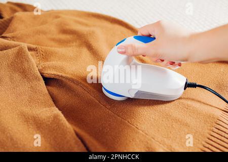 Frau, die zu Hause mit einem Fuzz-Rasierer Fusseln von der Kleidung entfernt. Nahaufnahme des kabelgebundenen elektrischen Handgeräts. Ich kümmere mich um die Kleidung. Haushaltsmaschine Stockfoto
