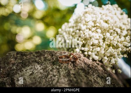 Zwei goldene Eheringe auf einem Stein, ein Blumenstrauß der Braut im Hintergrund Stockfoto
