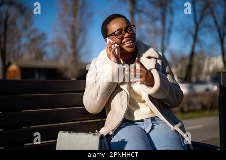 Außenporträt einer schwarzen Frau am sonnigen Tag. Sie sitzt auf einer Bank auf der Straße und telefoniert. Stockfoto