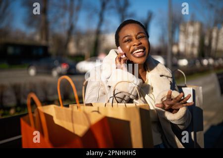 Außenporträt einer glücklichen schwarzen Frau. Sie sitzt nach dem Einkaufen auf der Bank auf der Straße. Stockfoto