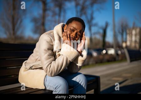 Außenporträt einer depressiven afroamerikanischen Frau am sonnigen Tag. Sie sitzt auf einer Bank auf der Straße. Stockfoto