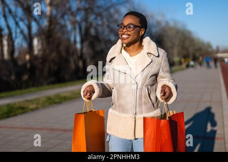Außenporträt einer glücklichen schwarzen Frau. Sie hält Einkaufstüten in der Hand und läuft auf der Straße herum. Stockfoto