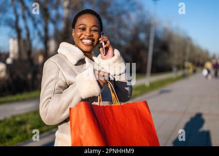 Außenporträt einer glücklichen schwarzen Frau. Sie hält Einkaufstüten in der Hand und telefoniert. Stockfoto