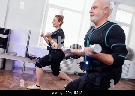 Ein paar lächelnde ältere Menschen in notarztanzügen, die im Fitnessraum mit Kurzhanteln trainieren. Grauhaariger Mann und dicke Frau trainieren mit persönlichem T-Shirt Stockfoto