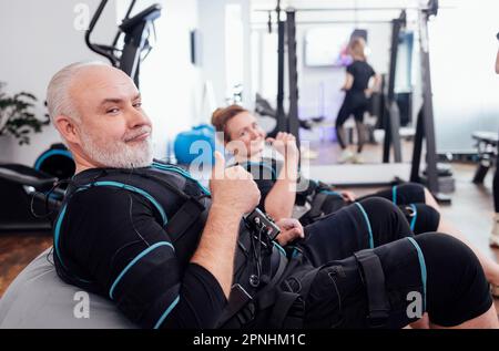 Ein älteres Paar sitzt auf Massagesesesseln und wartet auf das training im Fitnessraum oder Fitnessklub. Ältere grauhaarige Männer und Frauen in rettungssanitätern machen sich fertig Stockfoto