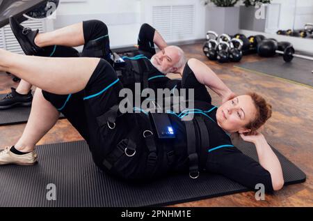 Sport ein paar ältere Menschen in ems-Anzügen trainieren im Fitnessraum oder Fitnessclub. Ein Mann mit grauem Bart und eine dicke Frau, die Bauchmuskeln auf dem Boden pumpt Stockfoto