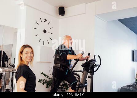 Persönliche Trainerin mit ihrem Klienten im rettungssanitäter-Anzug im Fitnessstudio. Der grauhaarige Sportler trainiert im Hintergrund auf einem Ellipsentrainer. Elektrik Stockfoto