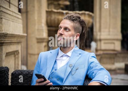 Ein junger erfolgreicher Mann im blauen Anzug mit einem Smartphone in der Hand sitzt in einem luxuriösen Straßenrestaurant Stockfoto