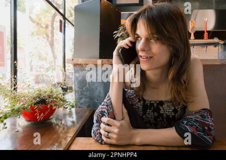 Wunderschöne junge transsexuelle latina-Frau argentinischer Herkunft, in einem Restaurant, das am Telefon sitzt, sich auf den Tisch lehnt Stockfoto