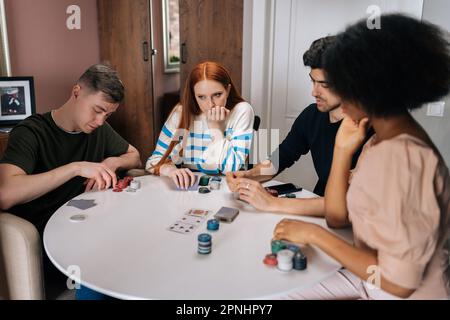 Porträt von pensiven, multiethnischen Freunden, die am Wochenende eine Party feiern und Pokerspiel im Wohnzimmer am Tisch spielen. Stockfoto