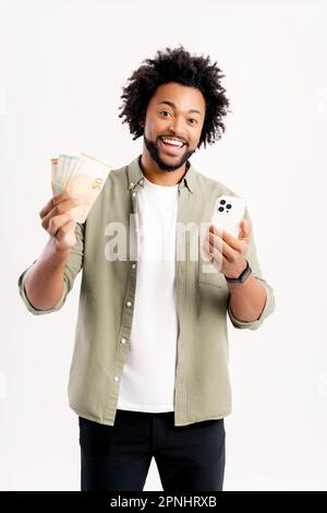 Überglücklich aufgeregter afroamerikanischer charismatischer Kerl mit Euro-Bargeld und Smartphone, fröhlicher Mann, der emotional auf Online-Sieg reagiert isoliert auf weißem Stand, Freelancer, die online verdienen Stockfoto