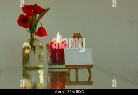 Eine hölzerne Staffelei steht neben einer Glasvase mit rotem Mohn und einer roten Kerze mit brennender Flamme auf einem Tisch. Einrichtung für ein gemütliches Zuhause. Zeichnung L Stockfoto