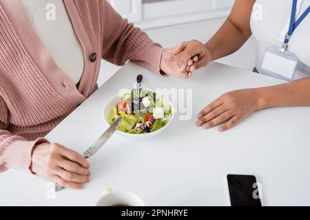 Draufsicht einer Seniorin, die Händchen hält, mit einer multiethnischen Pflegekraft neben dem Mittagessen auf dem Tisch Stockfoto