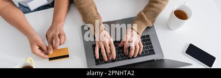 Draufsicht einer Seniorin, die auf einer Laptop-Tastatur in der Nähe einer multiethnischen Sozialarbeiterin tippt, mit Kreditkarte, Banner Stockfoto