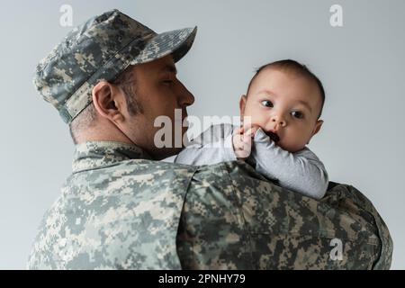 Fröhlicher Vater in Militäruniform und Mütze umarmt neugeborenen Sohn isoliert auf grau Stockfoto