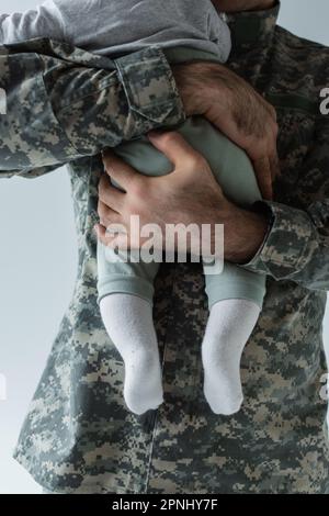Die Schnittansicht des Vaters in Militäruniform, der den Säuglingssohn umarmt, isoliert auf grau Stockfoto