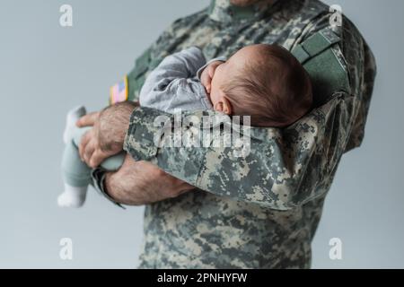 Schnittbild eines Soldaten in Uniform, der das Neugeborene auf grau isoliert hält Stockfoto