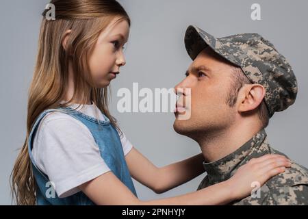 Trauriges Mädchen, das einen Soldaten in Militäruniform ansieht, während des Gedenkfeiertags weinend, isoliert auf Grau Stockfoto