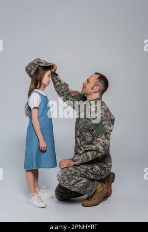 Glücklicher Soldat in Armeeuniform mit Militärmütze auf dem Kopf einer süßen Tochter auf Grau Stockfoto