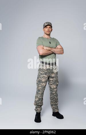 Ein Soldat in voller Länge in T-Shirt und Mütze, der während des Gedenkfeiertags mit zusammengefalteten Armen auf grau steht Stockfoto