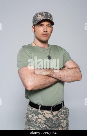 Soldat in T-Shirt und Mütze, der während des Gedenkfeiertags mit gefalteten Armen isoliert auf Grau stand Stockfoto