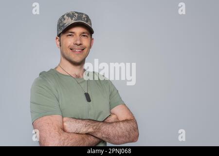 Glücklicher amerikanischer Soldat in T-Shirt und Militärmütze, der während des Gedenkfeiertags mit gefalteten Armen isoliert auf Grau stand Stockfoto