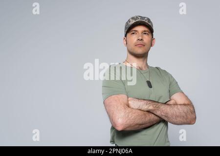 Militärmann in T-Shirt und Mütze mit gefalteten Armen während des Gedenkfeiertags isoliert auf Grau Stockfoto
