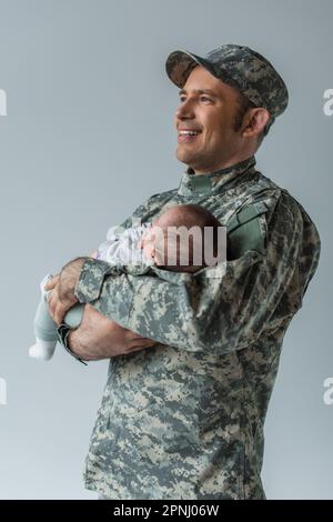 Fröhlicher Vater in Militäruniform und Mütze umarmt Neugeborenen isoliert auf grau Stockfoto