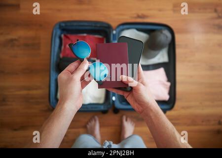 Koffer packen und Sommerabenteuer planen. Ein Mann, der ein Smartphone, einen Reisepass und eine Sonnenbrille in der Hand über dem offenen Gepäck hält. Stockfoto