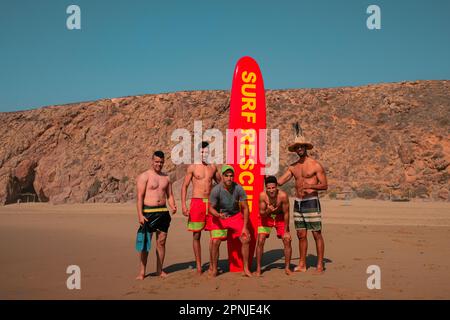 Fünf männliche Rettungsschwimmer am Aftas Beach posieren mit einem großen roten Surfbrett. Surf Rescue in gelben Buchstaben. Zerklüftete Landschaft in MIRLEFT, Marokko. Küstenstadt. Stockfoto