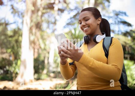 Glückliche brasilianische Studentin mit Smartphone und Rucksack, die im Park spaziert, auf den Bildschirm schaut und lächelt, freier Platz Stockfoto