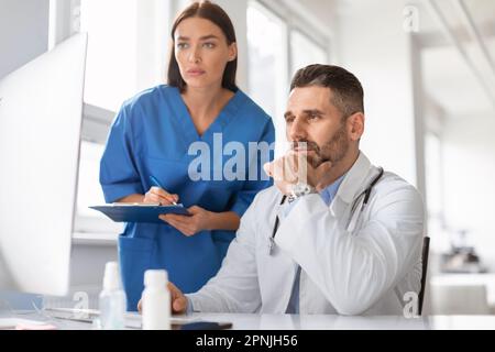 Fokussierte zwei Ärzte, die sich Computerbildschirme ansehen, die Krankheiten oder Diagnosen des Patienten diskutieren, in der Klinik zusammenarbeiten Stockfoto