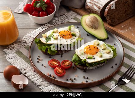 Toasts mit Avocado und Spiegeleiern auf dem Tisch. Gesundes Frühstück mit Gemüse und Kräutern. Spiegelei auf einem grauen Teller mit Orangensaft. Ich serviere auf dem Stockfoto