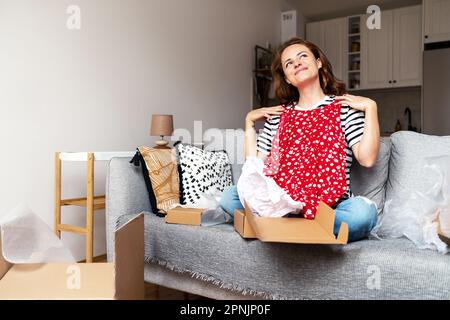Frau packt Pappkarton aus und kauft ihr neues Sommerkleid online im Internet-Shop. Stockfoto